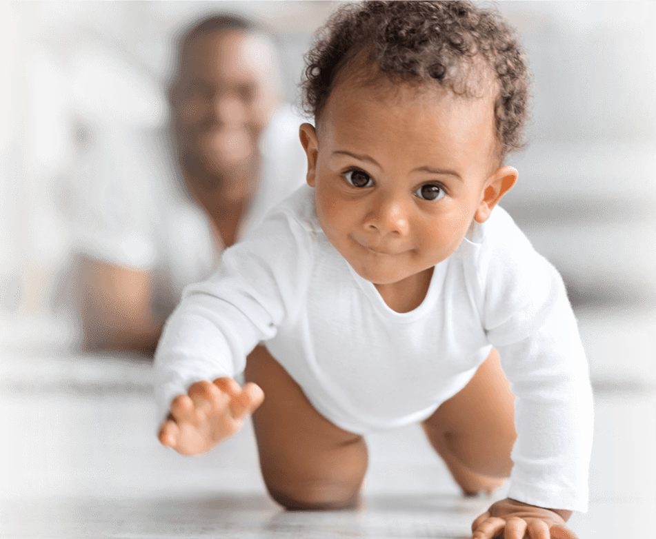 the baby crawls towards the camera on all fours. the background is blurred, but we can see the smiling father there