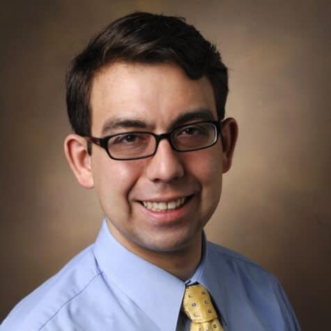 A headshot of a man with short, dark hair and glasses, smiling at the camera. He is wearing a light blue collared shirt and a yellow patterned tie. The background is a soft, mottled brown.