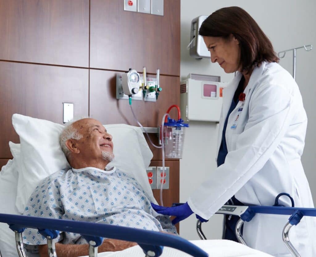 doctor standing beside elderly patient in bed. They are looking at each other smiling, and the doctor is touching the patient's arm in a reassuring way.