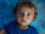 Young child with curly hair and striking blue eyes lying on a textured blue background, wearing a matching blue shirt.