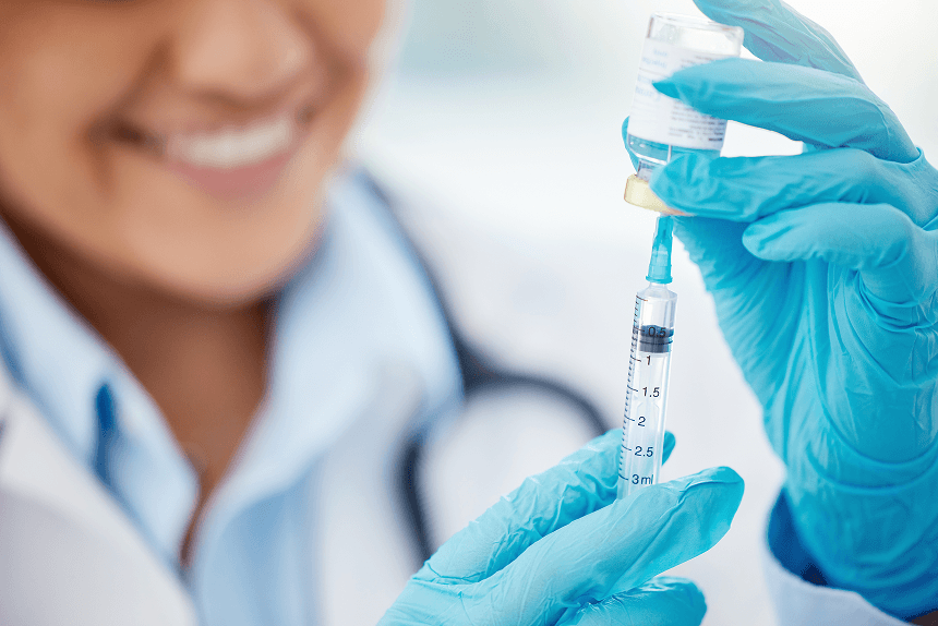 Smiling female healthcare professional in white coat and blue gloves holding a syringe and vaccine vial, preparing an injection.